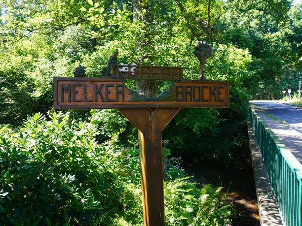 Ein Holzschild zeigt die Aufschrift "Melker Brücke" in einer grünen, bewaldeten Umgebung.
