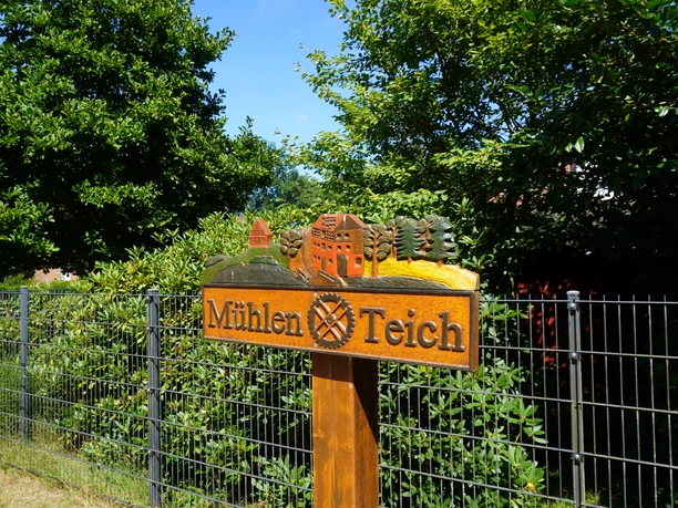 Ein Holzschild mit der Aufschrift "Mühlen Teich" steht vor grüner Hecke unter blauem Himmel.