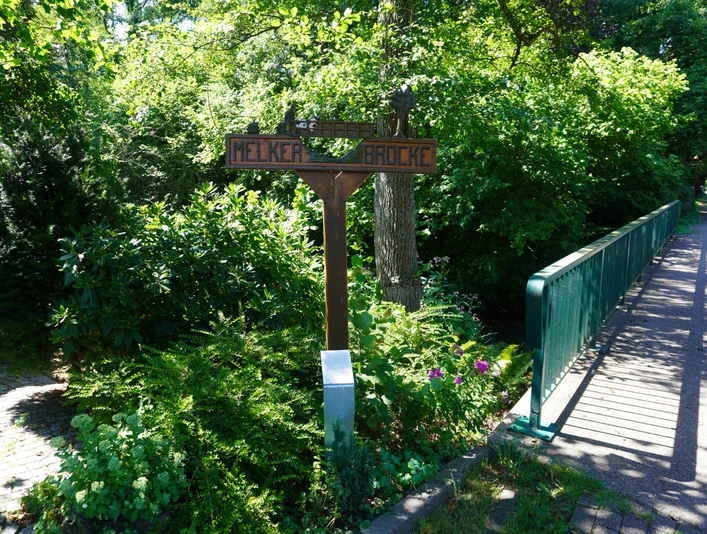 Ein hölzerner Wegweiser neben einer grünen Brücke, umgeben von dichtem, üppigem Laub.