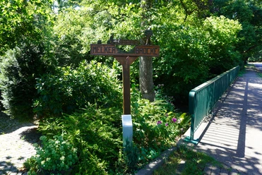 Eitzer Wegweiser Ein hölzerner Wegweiser neben einer grünen Brücke, umgeben von dichtem, üppigem Laub.