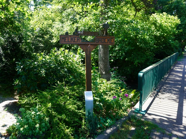 Eitzer Wegweiser Ein hölzerner Wegweiser neben einer grünen Brücke, umgeben von dichtem, üppigem Laub.
