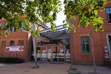 Eingang der Stadthalle Verden mit Glasfassade und rotem Ziegelbau, umrahmt von Kastanienblättern.
