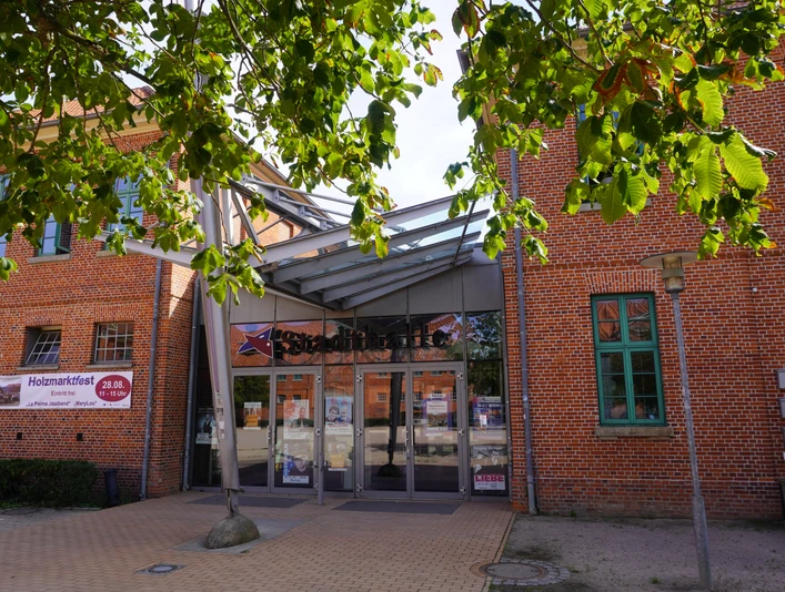 Eingang der Stadthalle Verden mit Glasfassade und rotem Ziegelbau, umrahmt von Kastanienblättern.