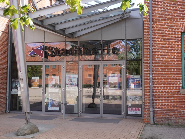 Eingang einer roten Backsteinhalle mit verglasten Türen und einem modernen Vordach aus Stahl und Glas.