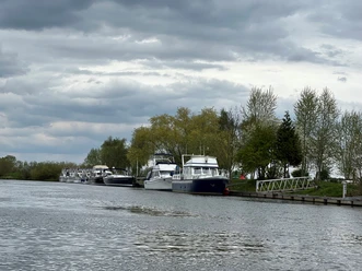 Motorboote liegen am ruhigen Flussufer, umgeben von Bäumen unter einem bewölkten Himmel.