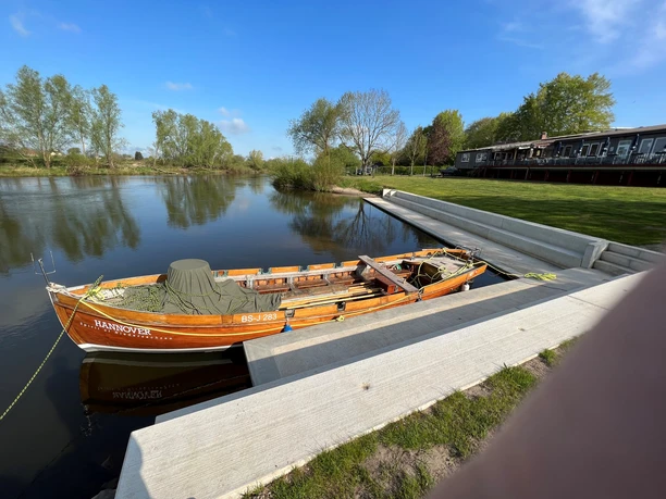 Ein traditionelles Holzboot mit der Aufschrift "Hannover" liegt an einem ruhigen Flussufer vertäut.