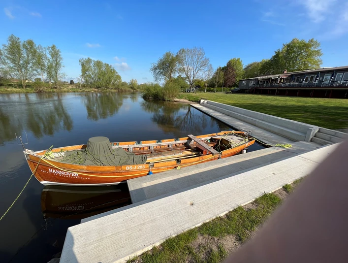 Wassersportverein Verden Ein traditionelles Holzboot mit der Aufschrift "Hannover" liegt an einem ruhigen Flussufer vertäut.