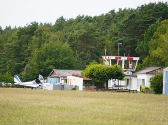 Flughafen Verden Scharnhorst Ein Sportflugzeug parkt auf einem grasbewachsenen Flugplatz vor einem kleinen weißen Flughafengebäude.