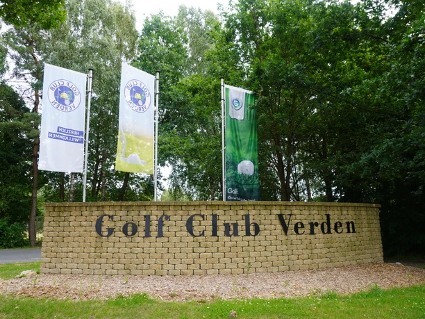 Entrance to the Verden Golf Club with three flags and a stone wall in green, secluded surroundings.