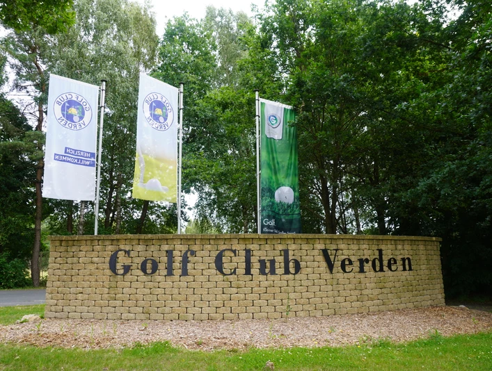 Eingang zum Golf Club Verden mit drei Fahnen und einer Steinmauer in grüner, lauschiger Umgebung.Entrance to the Verden Golf Club with three flags and a stone wall in green, secluded surroundings.Indgang til Verden Golf Club med tre flag og en stenmur i grønne, hyggelige omgivelser.Toegang tot de Verden Golf Club met drie vlaggen en een stenen muur in een groene, gezellige omgeving.