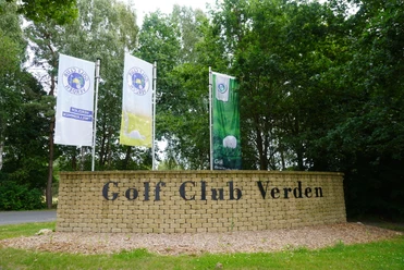Golfplatz Verden Eingang zum Golf Club Verden mit drei Fahnen und einer Steinmauer in grüner, lauschiger Umgebung.
