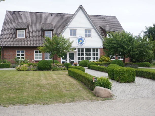 Bakstenen gebouw met verzorgde tuin voor de ingang van de golfbaan van Verden onder een grijze hemel.