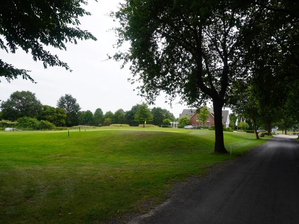 Groene fairways en bomen op een golfbaan in Verden, met een gebouw op de achtergrond, onder een bewolkte hemel.
