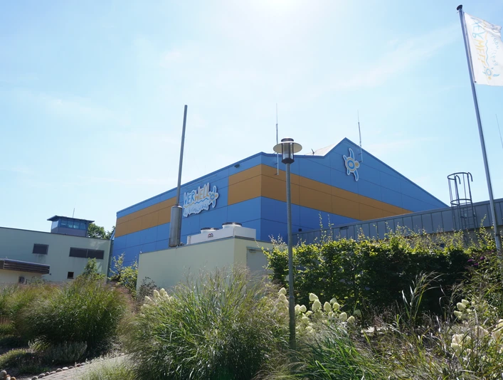 Wellnessbad mit markant blauer und oranger Fassade, umgeben von blühenden Gräsern bei klarem Himmel.