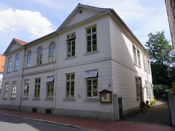 Wohnhaus Anita Augspurg Ein zweigeschossiges, weißes Wohngebäude mit Satteldach und vielen Fenstern an einer ruhigen Straße.