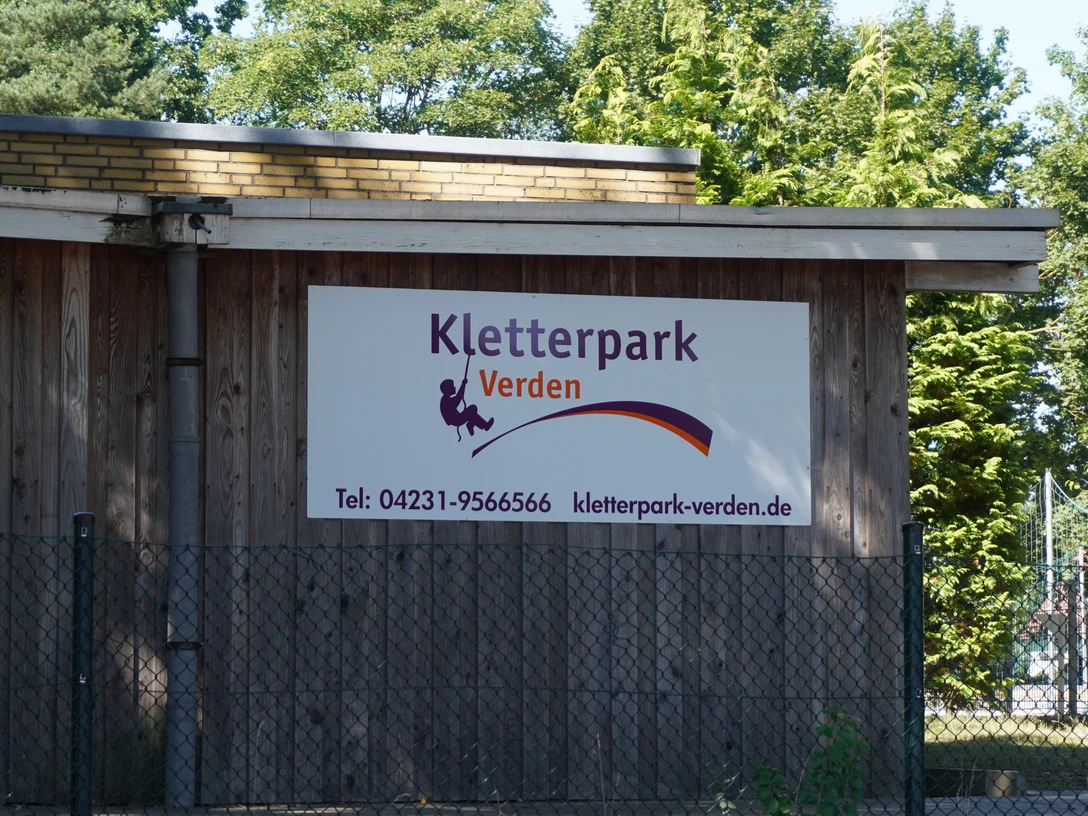 Kletterpark Verden Holzgebäude mit Schild des Kletterparks Verden, umgeben von Bäumen, bietet Abenteuer und Natur.Wooden building with sign of the Verden climbing park, surrounded by trees, offers adventure and nature.Træbygning med skilt til Verden klatrepark, omgivet af træer, byder på eventyr og natur.Houten gebouw met uithangbord van klimpark Verden, omgeven door bomen, biedt avontuur en natuur.
