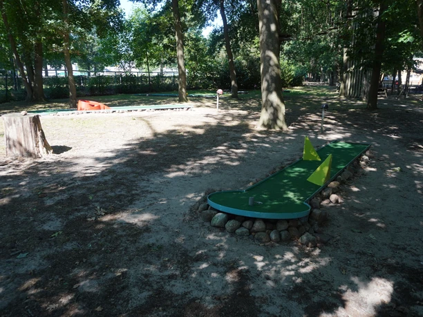 Minigolfbaan in de schaduw van hoge bomen in een bosrijke omgeving, met gele hindernissen en een natuurlijke omgeving.