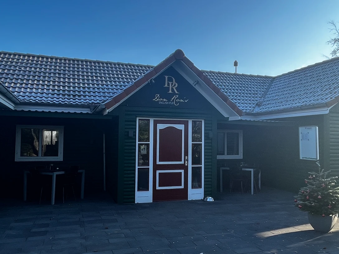 Don Ron's English Pub Gemütliches englisches Pub mit dunkelgrüner Fassade und roten Akzenten, umgeben von klarer Natur.