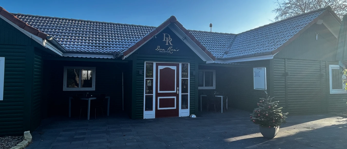 Don Ron's English Pub Gemütliches englisches Pub mit dunkelgrüner Fassade und roten Akzenten, umgeben von klarer Natur.