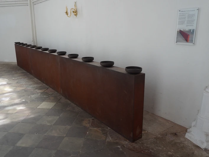 Die Skulptur zeigt zehn dunkle Schalen auf einem langen, rechteckigen Metallblock in einem Innenraum.