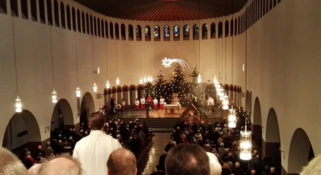 Piuskirche_Wiedenbrück.jpg Eine festliche Weihnachtsmesse in einer Kirche mit großen Kronleuchtern, geschmückten Tannenbäumen und Menschen.