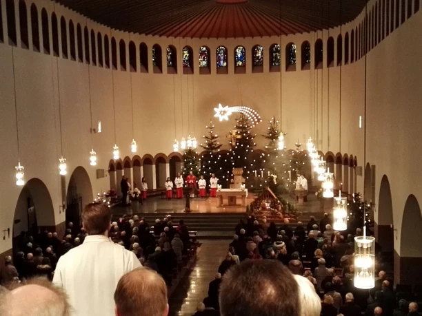 Piuskirche_Wiedenbrück.jpg Eine festliche Weihnachtsmesse in einer Kirche mit großen Kronleuchtern, geschmückten Tannenbäumen und Menschen.