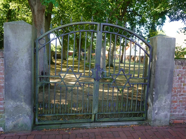 A historic gate adorned with Stars of David leads to a tree-lined cemetery.