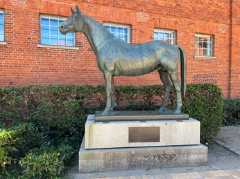 Tempelhüter Bronzestatue eines Pferdes, des Tempelhüters, vor einer roten Backsteinmauer mit Fenstern.