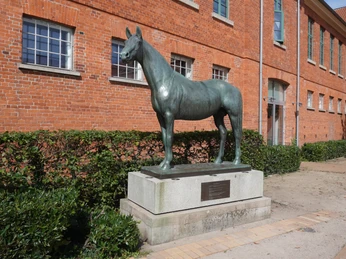 Tempelhüter Bronzene Pferdeskulptur "Tempelhüter" auf Sockel vor einer roten Backsteinfassade.