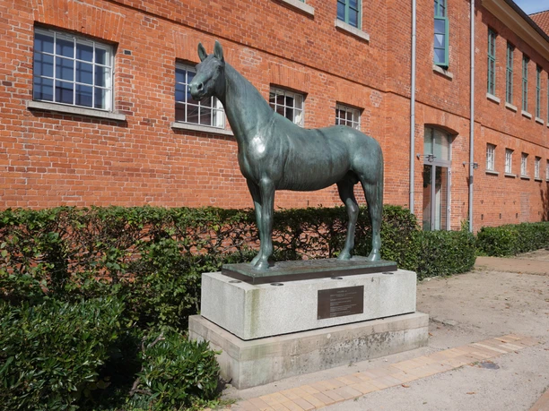 Tempelhüter Bronzene Pferdeskulptur "Tempelhüter" auf Sockel vor einer roten Backsteinfassade.