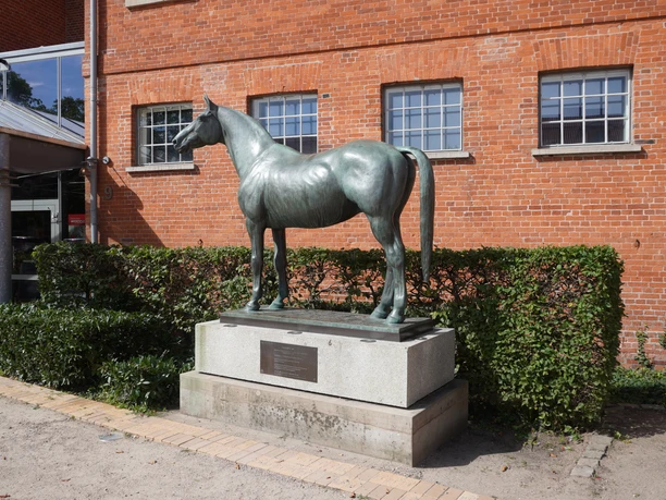 Die Bronzestatue eines Pferdes steht auf einem Sockel vor einer roten Backsteinfassade.
