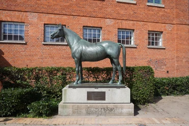 Bronzestatue eines Pferdes namens Tempelhüter vor roter Backsteinfassade, umgeben von Büschen.
