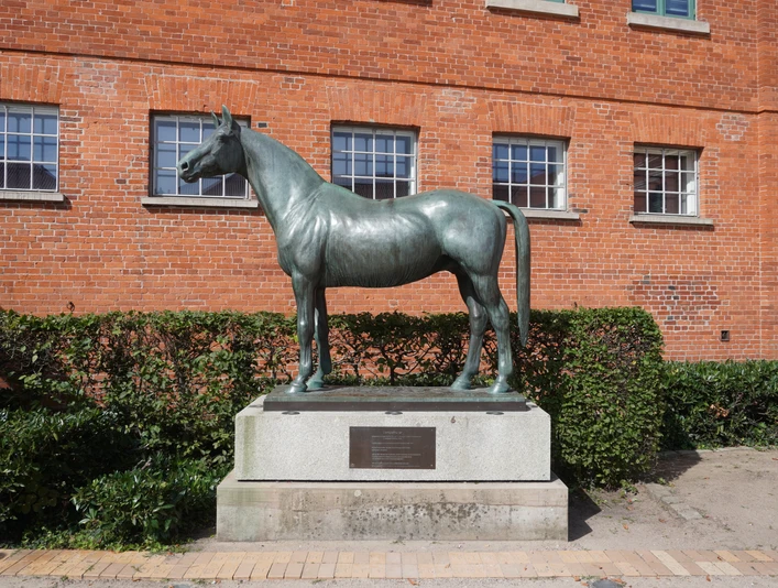 Tempelhüter Bronzestatue eines Pferdes namens Tempelhüter vor roter Backsteinfassade, umgeben von Büschen.