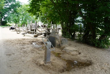 Naturbelassener Spielplatz in Verden mit Holzgeräten zum Klettern, Sandfläche und umliegenden Bäumen.