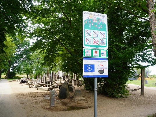 Spielplatz mit Holzkonstruktionen und Infotafel im Allerpark Verden, umgeben von Bäumen.