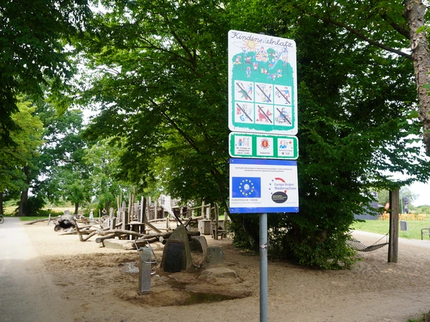 Spielplatz mit Holzkonstruktionen und Infotafel im Allerpark Verden, umgeben von Bäumen.