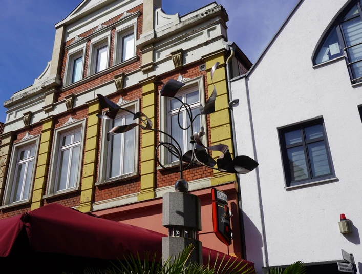 Moderne Kunstinstallation "Wachstumssäule" vor historischem Gebäude mit markanten Fenstern und Geländer.