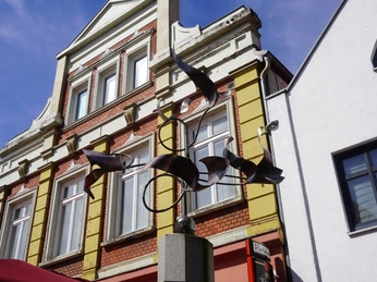 Metallskulptur mit geschwungenen Formen thront auf hohem Sockel vor historischem Gebäude.