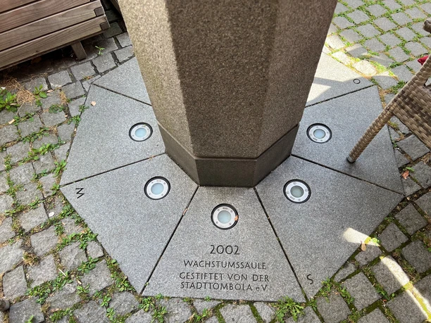 Wachstumssäule Steingrauer Sockel mit integrierten Leuchten umgibt 2002 gestiftete Wachstumssäule auf Pflastersteinen.