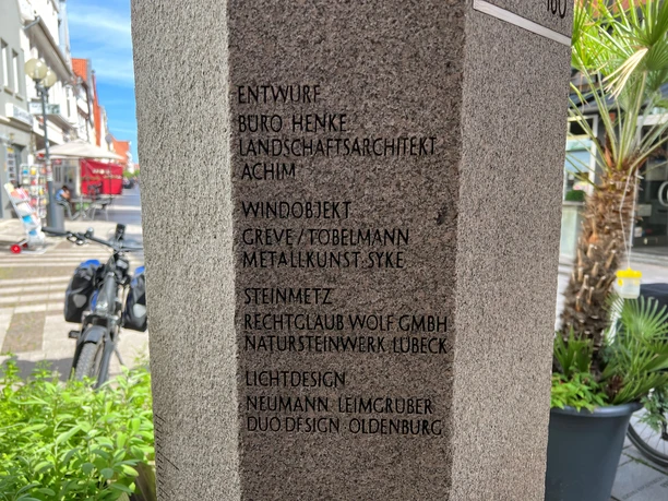Eine steinerne Säule im urbanen Raum mit eingravierten Namen und Kunstwerken umgeben von Fahrrädern.