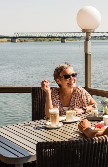 Eine Frau und ein Mann sitzen auf der Terasse im "Cafe von Herzen" in Lauenburg. Die Sonne scheint und sie blicken direkt auf die Elbe während sie Kaffee und Kuchen genießen.