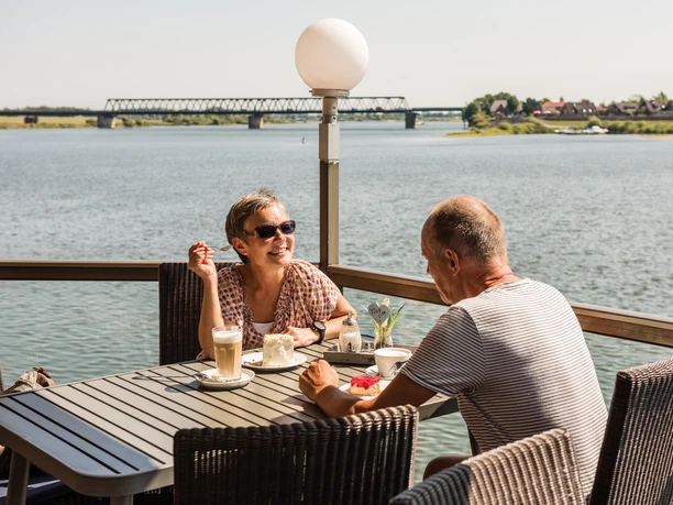 Eine Frau und ein Mann sitzen auf der Terasse im "Cafe von Herzen" in Lauenburg. Die Sonne scheint und sie blicken direkt auf die Elbe während sie Kaffee und Kuchen genießen.