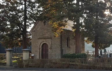 Kreuzkapelle in Kürten-Olpe Eine kleine, historische Kapelle aus Stein, umgeben von großen Bäumen, auf einem Friedhof im Sonnenschein.