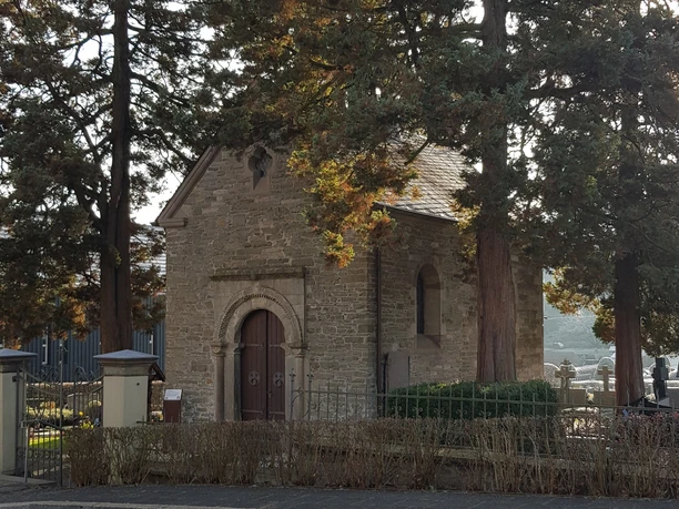 Kreuzkapelle in Kürten-Olpe Eine kleine, historische Kapelle aus Stein, umgeben von großen Bäumen, auf einem Friedhof im Sonnenschein.