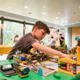 FIRST_LEGO_League_HNF.jpg Ein Junge konzentriert sich beim Aufbau eines LEGO-Projekts, umgeben von bunten LEGO-Modellen.