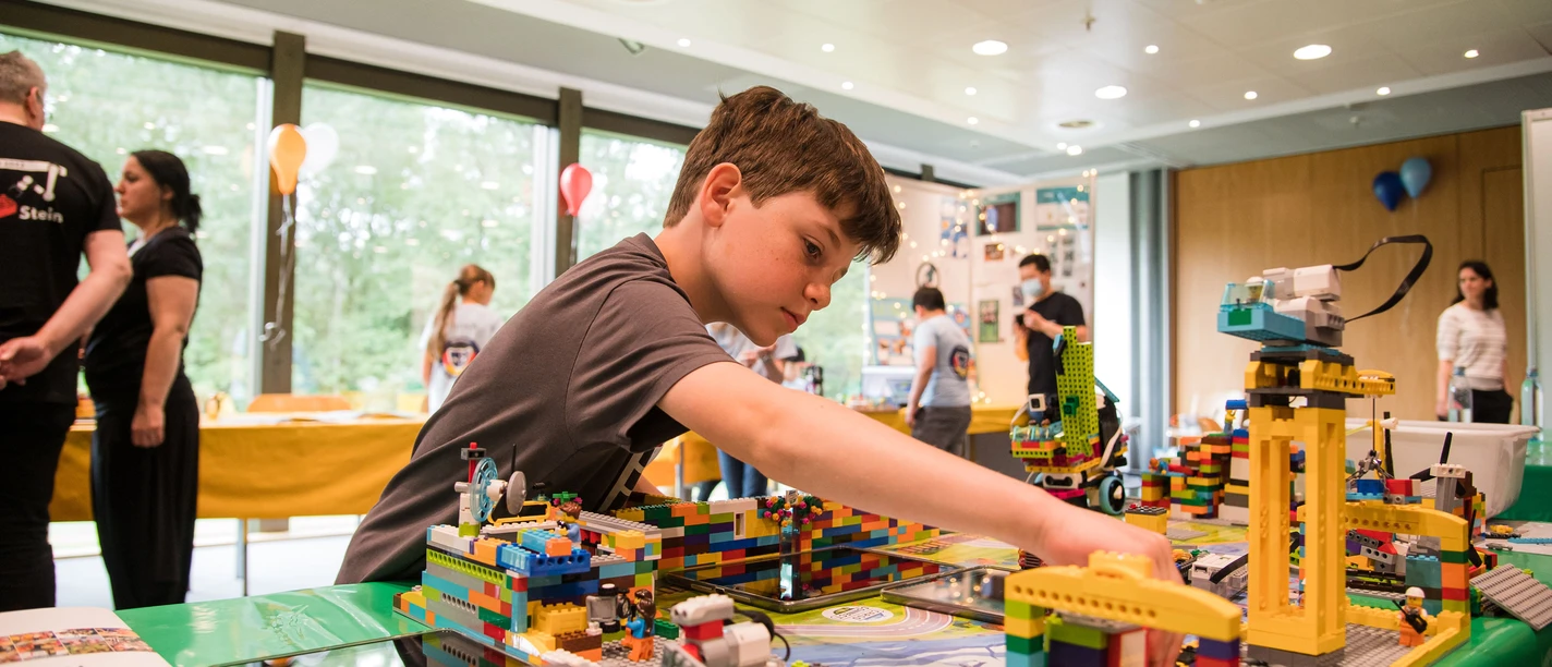 FIRST_LEGO_League_HNF.jpg Ein Junge konzentriert sich beim Aufbau eines LEGO-Projekts, umgeben von bunten LEGO-Modellen.