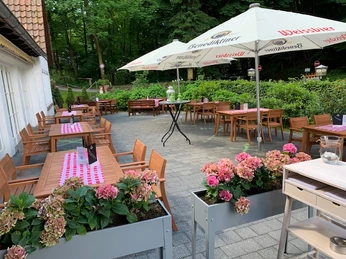 Hirschsprung_Terasse_2.jpg Einladende Außenterrasse mit Holztischen, umgeben von Blumen und Bäumen, geschützt durch Sonnenschirme.