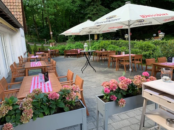 Hirschsprung_Terasse_2.jpg Einladende Außenterrasse mit Holztischen, umgeben von Blumen und Bäumen, geschützt durch Sonnenschirme.