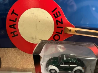 Ein altes Polizeischild mit einem Miniaturmodell eines grünen VW Käfer Polizeiautos im Vordergrund.
