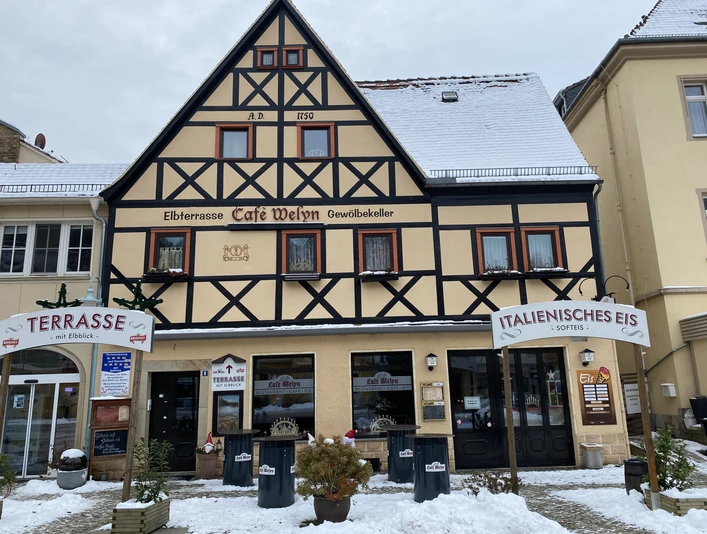 Café Welyn Fachwerkhaus mit Café und Eisdiele, beschneit, mit Schildern "Terrasse" und "Italienisches Eis", umgeben von winterlicher Atmosphäre.Half-timbered house with café and ice cream parlor, snow-covered, with signs "Terrace" and "Italian ice cream", surrounded by a wintry atmosphere.Polodřevěný dům s kavárnou a zmrzlinárnou, zasněžený, s nápisy "Terasa" a "Italská zmrzlina", obklopený zimní atmosférou.Dom z muru pruskiego z kawiarnią i lodziarnią, pokryty śniegiem, z napisami "Taras" i "Lody włoskie", otoczony zimową atmosferą.Vakwerkhuis met café en ijssalon, besneeuwd, met bordjes "Terras" en "Italiaans ijs", omgeven door een winterse sfeer.Casa a graticcio con bar e gelateria, innevata, con insegne "Terrazza" e "Gelato italiano", immersa in un'atmosfera invernale.